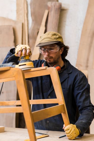 Man wearing safety eyeglasses working with orbital sanders on wooden furnitur