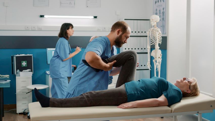 Physician doing legs raise exercise to stretch legs for old patient with mechanical disorders in a rehabilitation cabinet. An osteopathy specialist is doing a physical therapy procedure to cure a woman.
