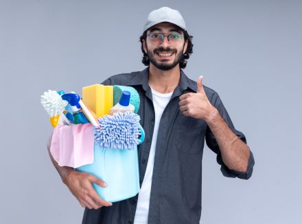 Smiling young handsome cleaning guy wearing t-shirt and cap holding bucket of cleaning tools showing thumb up isolated on white wall