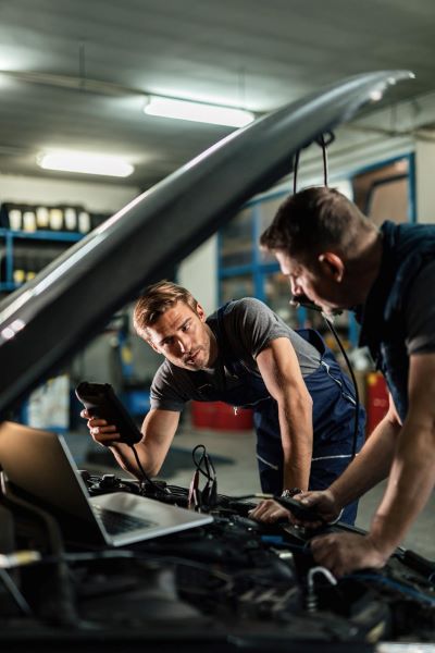 Young car repairman talking with his coworker while checking engine performance with diagnostic tools in auto repair shop